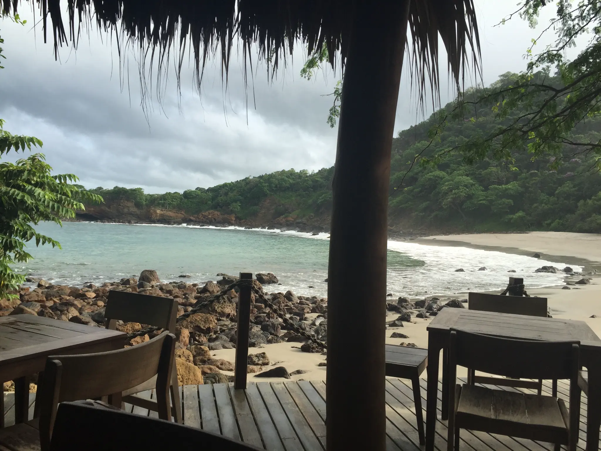Beachside restaurant at Aqua Resort Nicaragua — thatched palapa, wooden tables, turquoise cove, white sand, and jungle cliffs