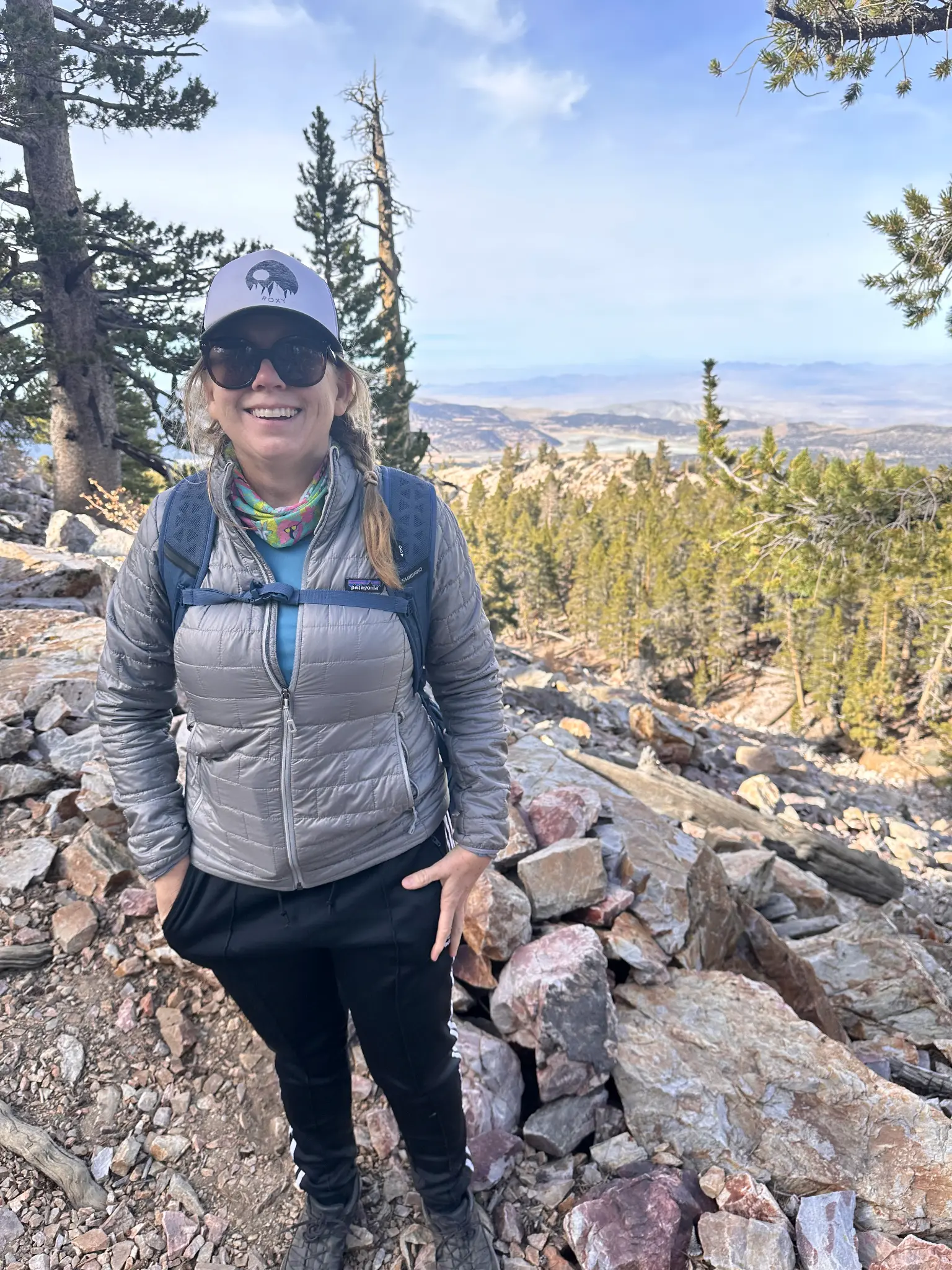 Kristen at the summit of Sugarloaf Mountain, smiling in a Patagonia jacket with a panoramic view of the Big Bear Valley and high desert stretching to the horizon