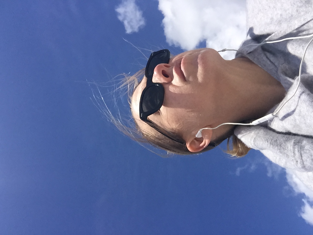 Kristen looking up at a bright blue sky with earbuds in, wind in her hair โ taken during a walk while waiting